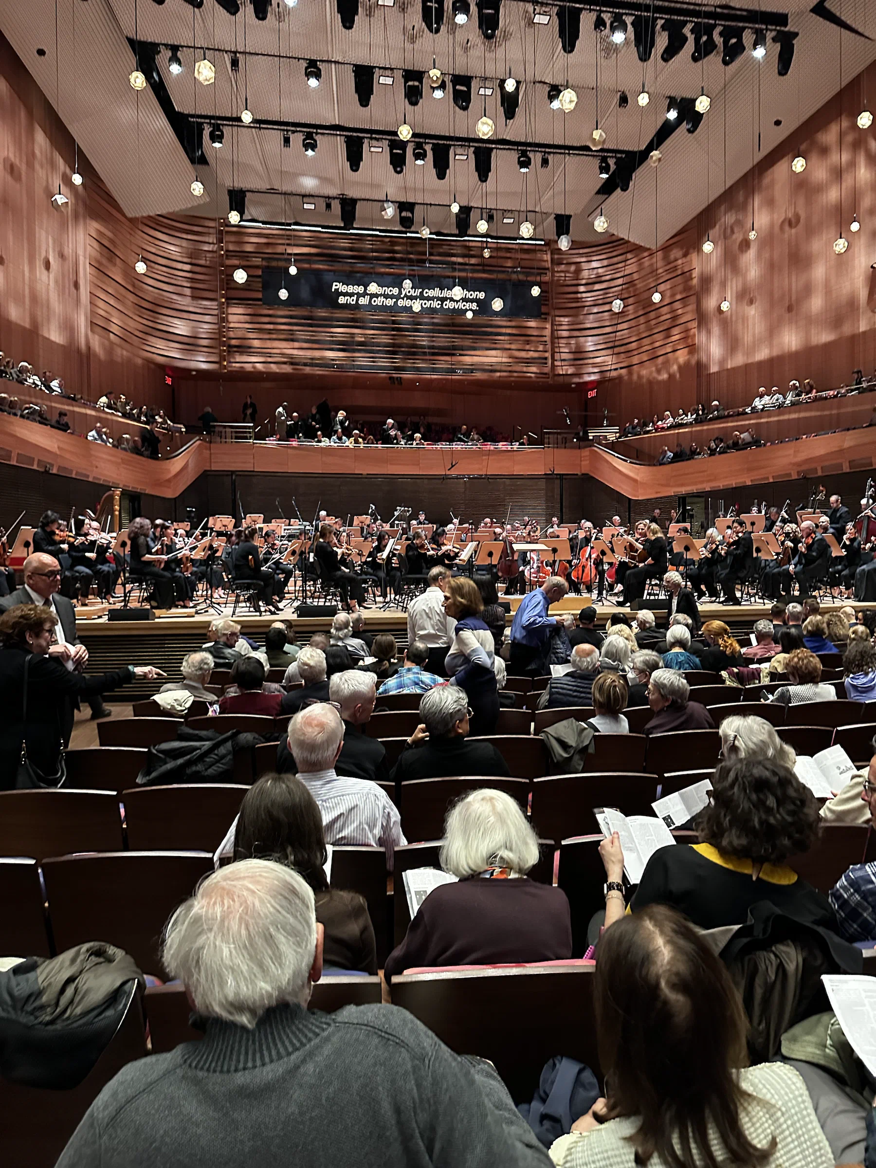 An orchestra performs in a large wood-paneled concert hall viewed from the audience.