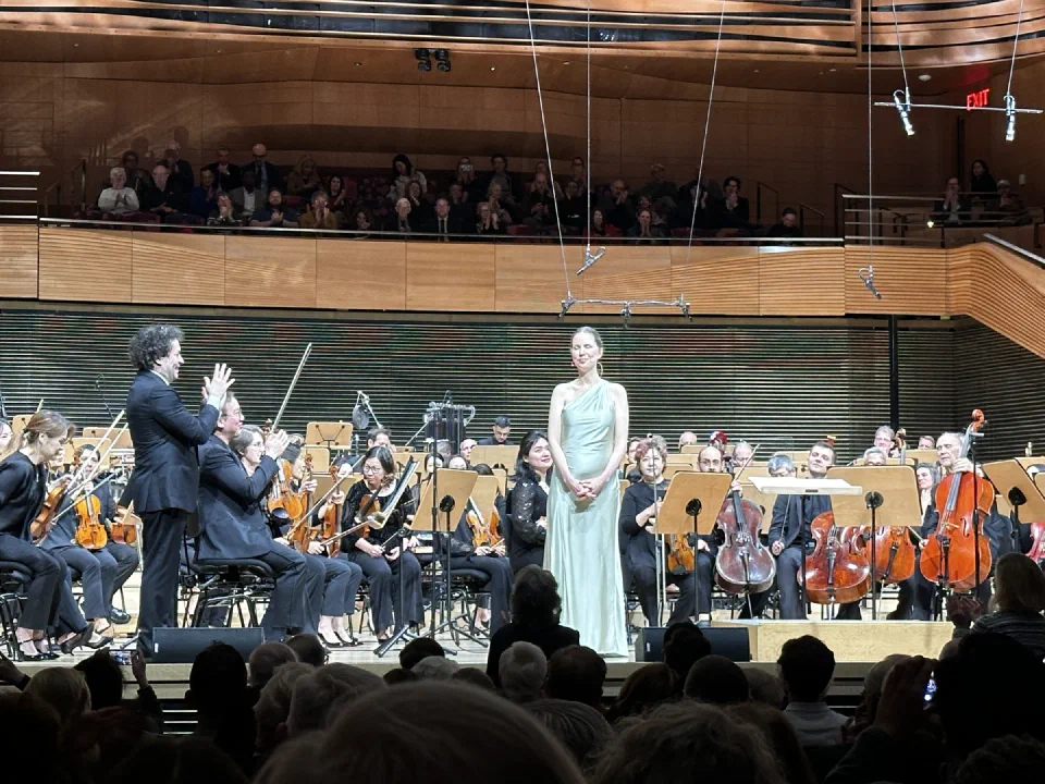 A singer performs before an orchestra in a wood-paneled concert hall.