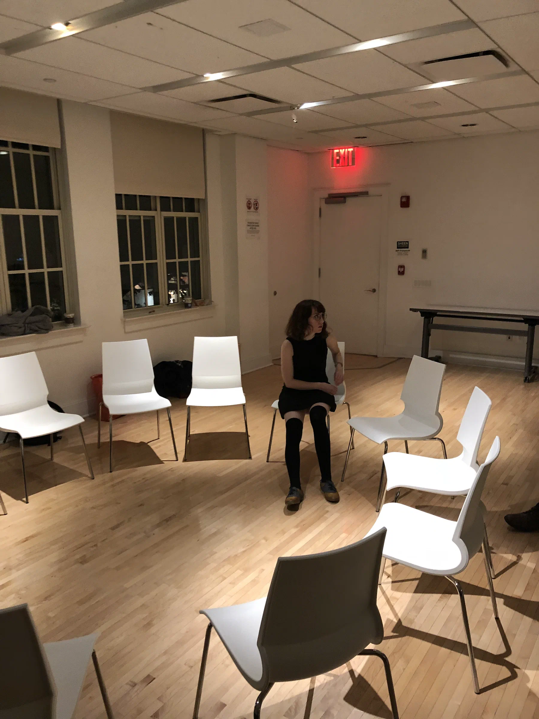 A woman sits alone within a circle of empty chairs in a plain meeting room.