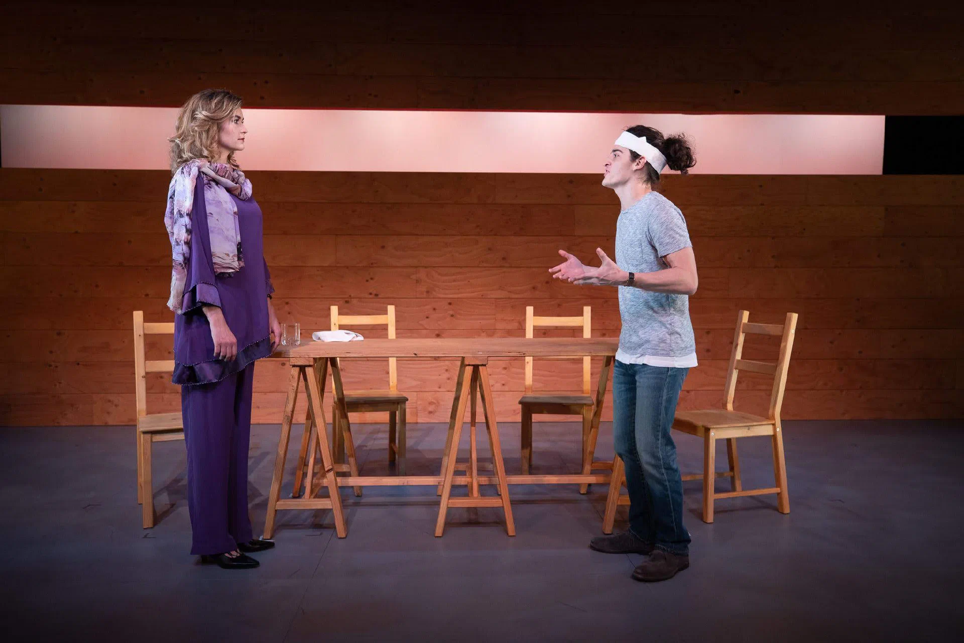 Two women face one another beside a loose arrangement of wooden chairs.