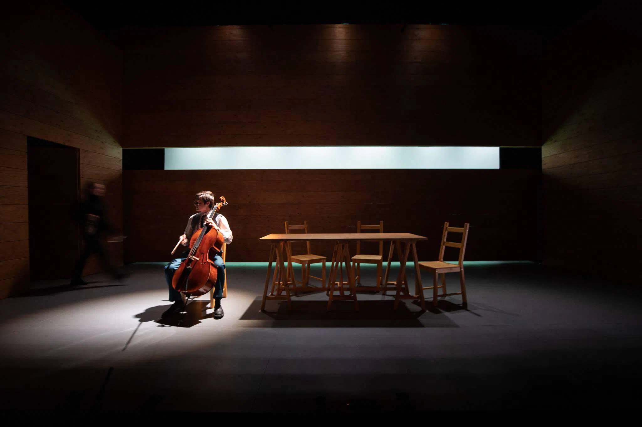 A cellist and performer occupy a dark stage divided by a band of white light.