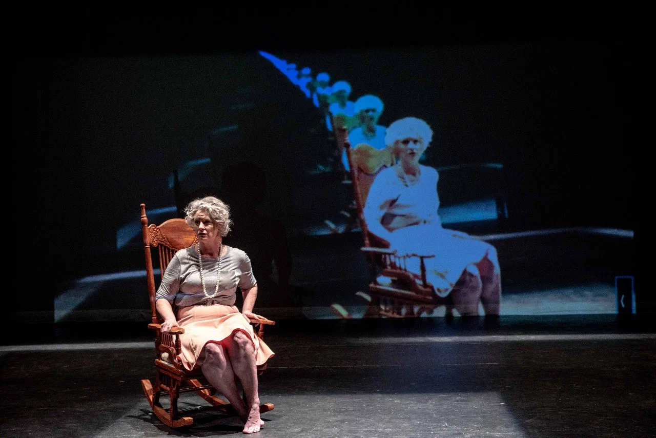 A seated older woman faces a large projected image of herself in a rocking chair.