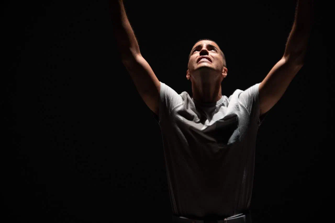 A lone performer reaches both arms upward against a black background.