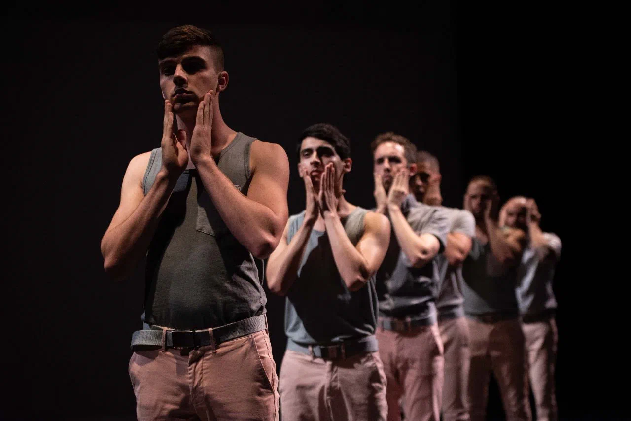 A line of performers holds their hands near their faces beneath focused stage light.