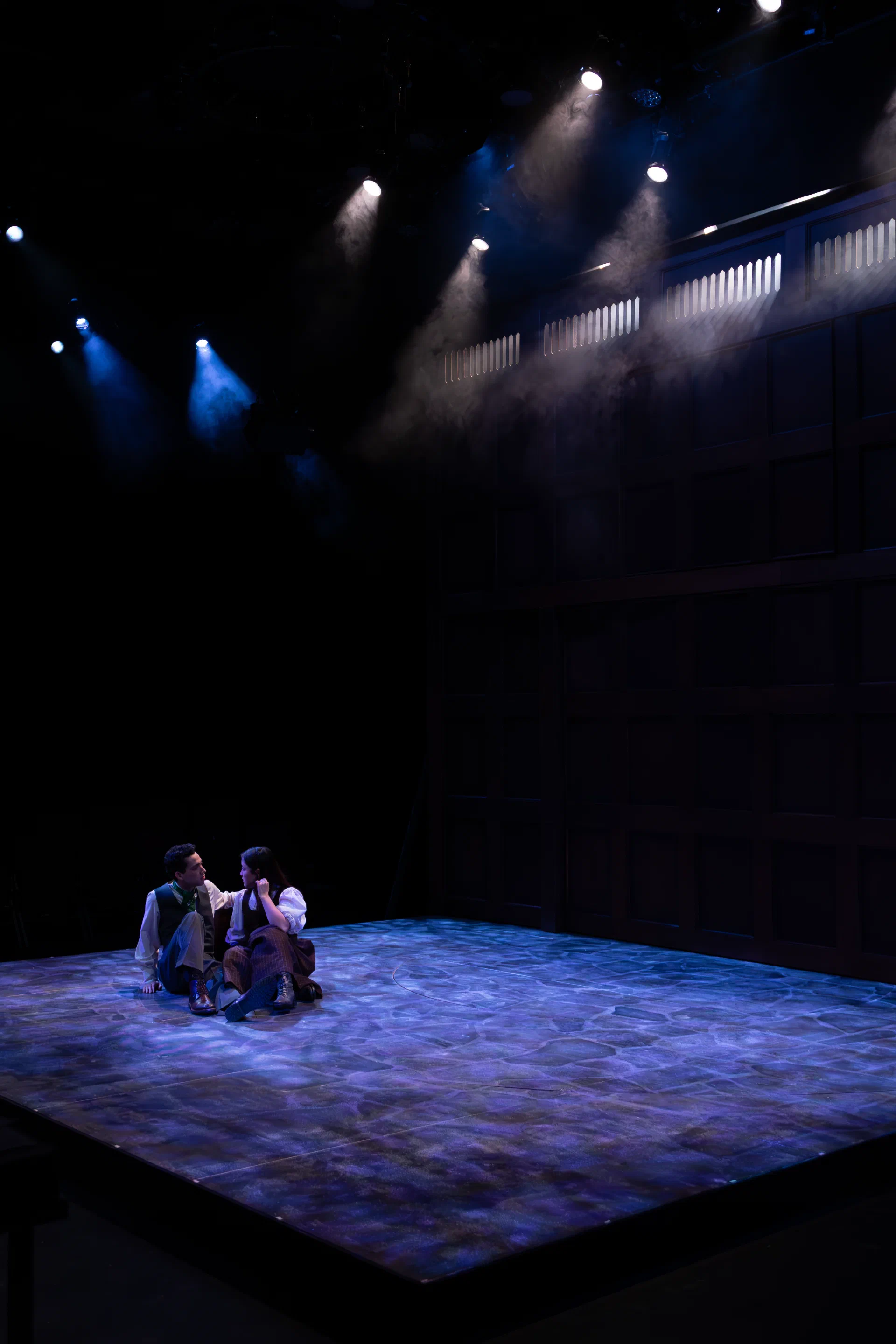 Two performers sit close together in a small pool of light on a dark stage.