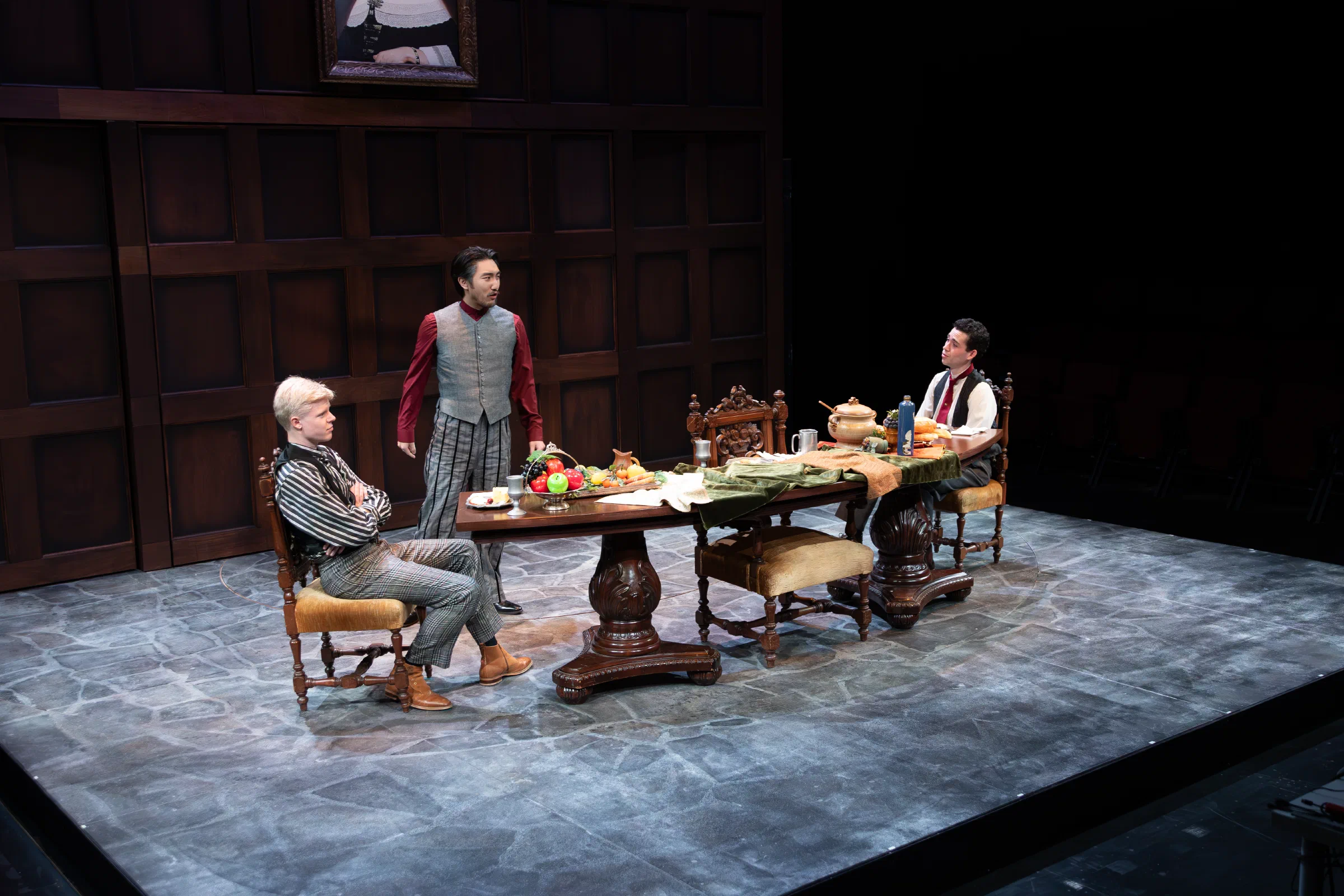 Three performers gather around a laden dining table on a dark wood-paneled set.