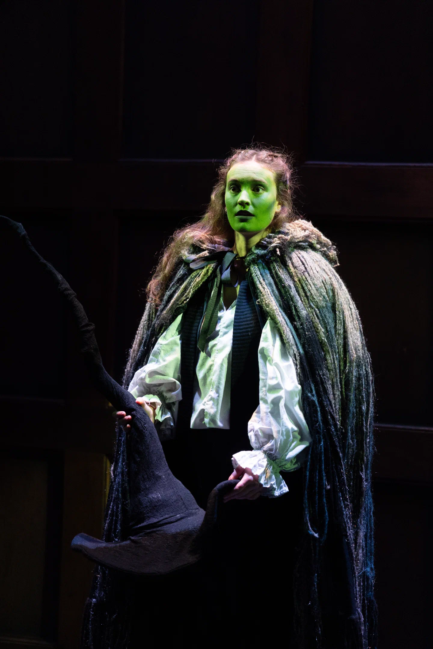 A performer with a green-painted face wears a dark cloak under focused stage light.