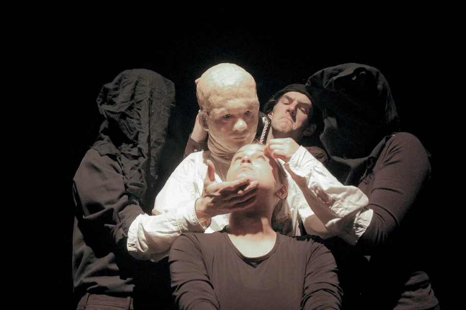 Three performers cluster around a seated figure in a starkly lit scene.